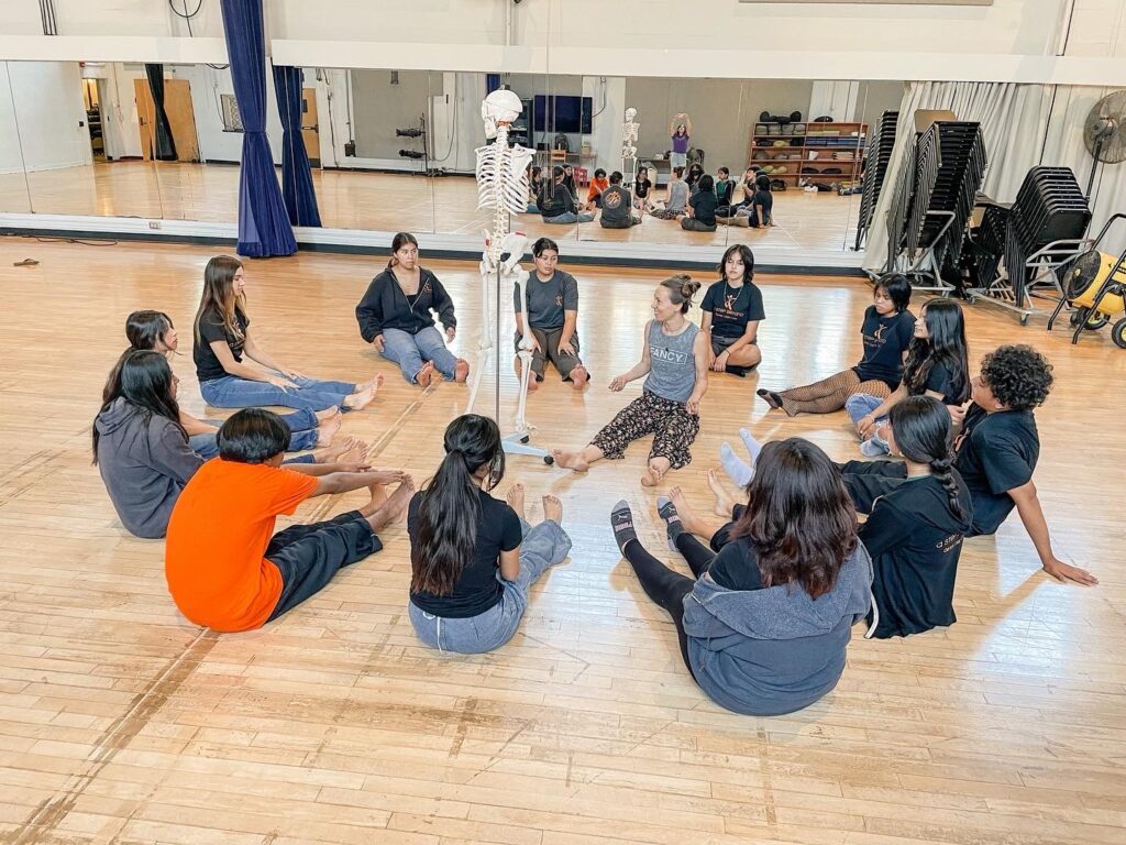 Students sitting in a circle with a dance instructor around a fake skeleton in a dance studio